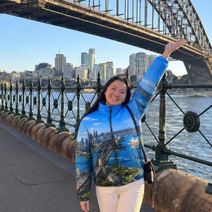 Könnte beinhalten: Eine blaue Steppjacke mit einem Fotodruck der Sydney Harbour Bridge und der Skyline der Stadt, ausgestattet mit einem hohen Kragen, einem durchgehenden Frontreißverschluss und langen Ärmeln, kombiniert mit einer weißen Hose.