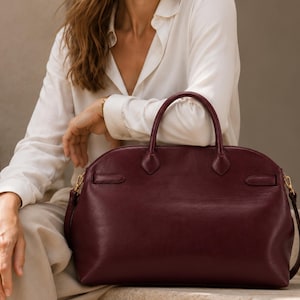 Könnte beinhalten: Eine tiefburgunderrote Ledertasche mit abgerundeter Oberseite, zwei stabilen Tragegriffen und goldfarbenen Metallbeschlägen für einen Schulterriemen. Die Tasche hat eine glatte, strukturierte Oberfläche mit einem horizontalen Riemendetail.