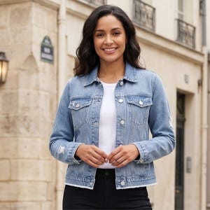 Puede incluir: Chaqueta vaquera azul claro con cuello clásico, cierre de botones y detalles desgastados. La chaqueta se lleva sobre una camiseta blanca y combinada con pantalones negros. La modelo sonríe frente a un edificio.