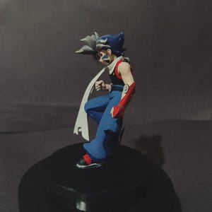 May include: A figurine of a character with blue hair and a white cape. The figure is wearing blue trousers, a red and black top, and red arm guards. The figure is posed in a dynamic stance on a black base.