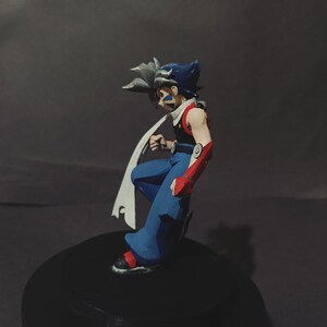 May include: A detailed figurine of an anime character in a dynamic pose. The figure features blue trousers, a red and black top, and a white scarf. The character has spiky hair with blue and grey tones, and is standing on a black base.