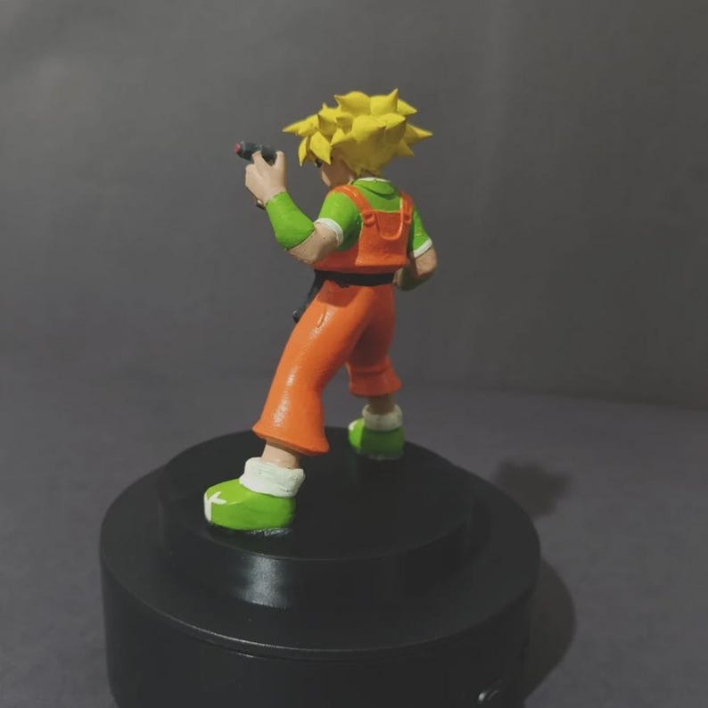 May include: A small figurine of a character with spiky yellow hair, wearing a green short-sleeved shirt under orange overalls, and green shoes. The character holds a small black object in one hand and stands on a circular black base.