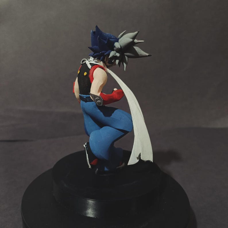 May include: A handmade figurine of a character in a dynamic pose. The figure has blue trousers, a black waistcoat with yellow details, red arm guards, and a white scarf. The hair is a mix of blue and grey. The figurine is on a black base.