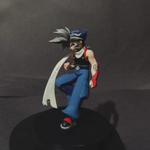 May include: A detailed figurine of an anime character in a dynamic pose. The figure has blue and grey spiky hair, blue trousers, a black top, and a white scarf. Red accents are visible on the arms and shoes. The figure is standing on a black base.