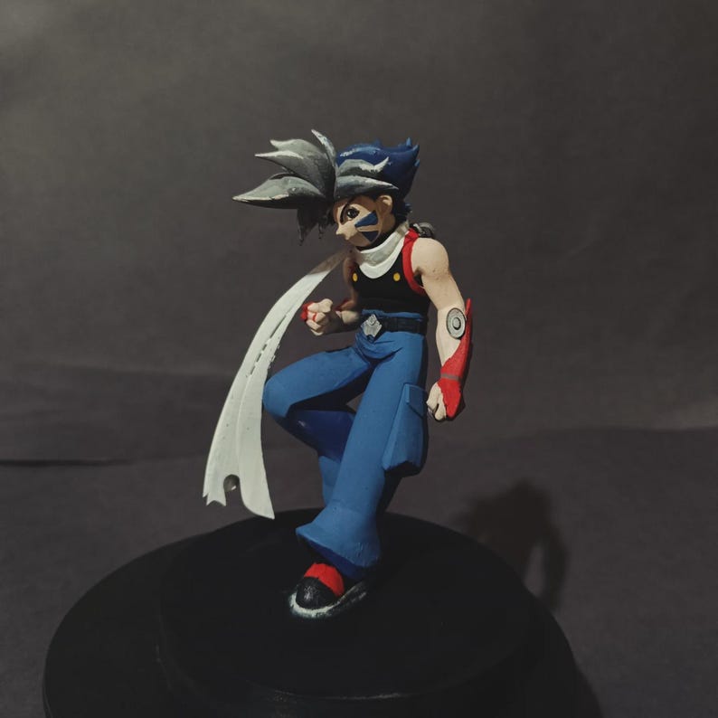 May include: A detailed figurine of an anime character in a dynamic pose. The figure has blue and grey spiky hair, blue trousers, a black top, and a white scarf. Red accents are visible on the arms and shoes. The figure is standing on a black base.