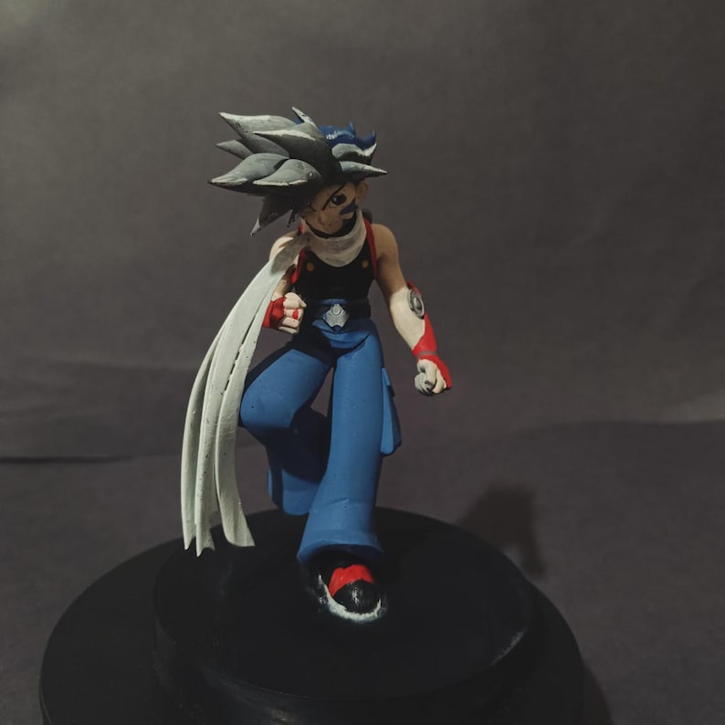 May include: A figurine of a character with spiky grey hair, wearing a black top, blue trousers, and red and white arm sleeves. The figure has a white scarf and is posed dynamically on a black base.