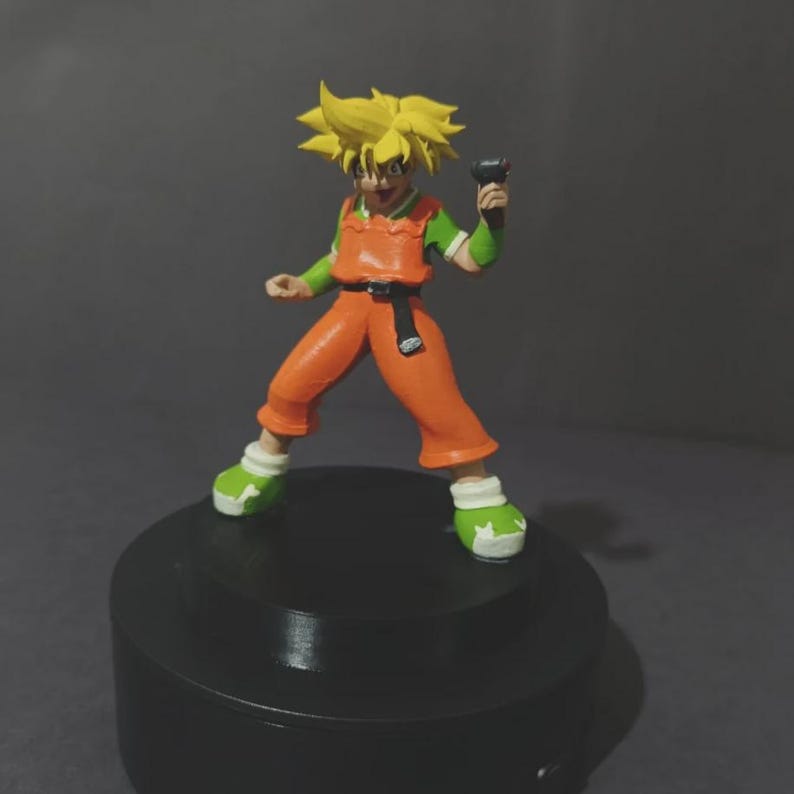 May include: A small figurine of a spiky-haired character with yellow hair, wearing an orange jumpsuit with green sleeves, a black belt, and green and white shoes. The character holds a small dark object in one hand and stands on a black base.