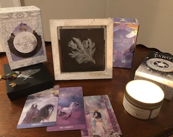 Può includere: Un piano da tavolo con una varietà di mazzi di carte da tarocchi, tra cui The Moonchild Tarot, The Starchild Tarot e un mazzo in bianco e nero. C'è anche una piccola statuetta di uccello, una cornice in legno bianco con un'immagine in bianco e nero di un cardo e una candela accesa.