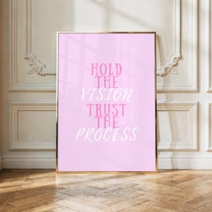 May include: A rectangular wall art print in a gold frame featuring a light pink background with the text HOLD THE VISION in pink block letters and TRUST THE PROCESS in white cursive script, centered on the surface of the print.