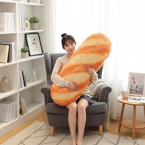 Puede incluir: Una gran almohada de baguette de felpa en tonos naranja y crema. La almohada tiene la forma de una barra de pan, con costuras detalladas para imitar la textura de una baguette. La sostiene una persona sentada en un sillón gris.
