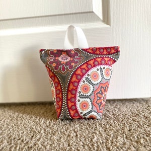May include: A red, gray, and white patterned doorstop with a white fabric handle. The doorstop is sitting on a beige carpet.