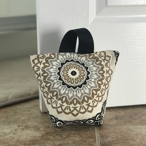 May include: A beige and black doorstop with a mandala pattern. The doorstop has a black fabric handle.