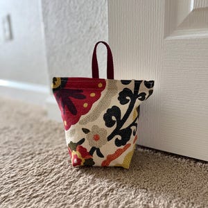 May include: A fabric doorstop with a colorful floral pattern in red, yellow, black, and gray on a cream background. It has a burgundy handle and is sitting on a beige carpet.