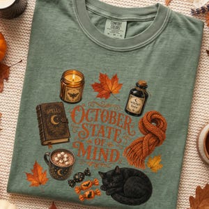 May include: A moss green t-shirt featuring a graphic print with the text "OCTOBER STATE OF MIND" surrounded by illustrations of a candle, a spell book, a maple leaf, a bottle labeled "witches brew", a knit scarf, a mug of cocoa, and a black cat.