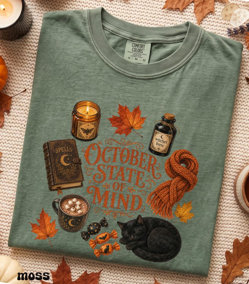 May include: A moss green t-shirt featuring a graphic print with the text "OCTOBER STATE OF MIND" surrounded by illustrations of a candle, a spell book, a maple leaf, a bottle labeled "witches brew", a knit scarf, a mug of cocoa, and a black cat.