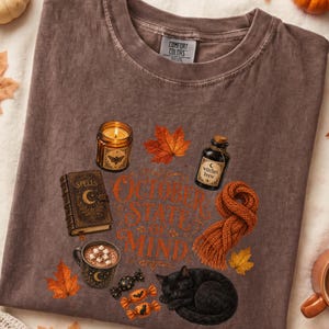 May include: A brown t-shirt featuring a graphic print with the text OCTOBER STATE OF MIND, a lit candle, a spell book, a bottle labeled Witches Brew, a mug of hot cocoa with marshmallows, a knitted orange scarf, a black cat, and autumn leaves.