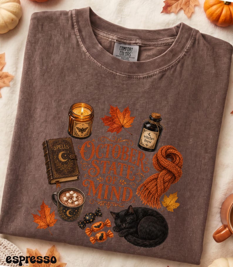 May include: A brown t-shirt featuring a graphic print with the text OCTOBER STATE OF MIND, a lit candle, a spell book, a bottle labeled Witches Brew, a mug of hot cocoa with marshmallows, a knitted orange scarf, a black cat, and autumn leaves.