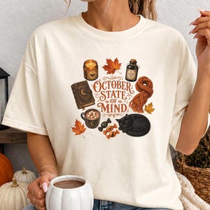 May include: An ivory short-sleeve t-shirt featuring a graphic print with the text "OCTOBER STATE OF MIND" surrounded by illustrations of a lit candle, a spell book, a mug of hot cocoa, a black cat, a knitted scarf, autumn leaves, and candies.