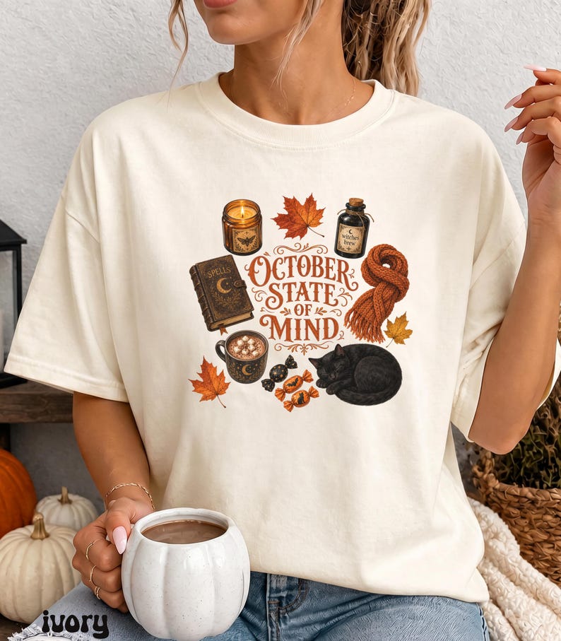 May include: An ivory short-sleeve t-shirt featuring a graphic print with the text "OCTOBER STATE OF MIND" surrounded by illustrations of a lit candle, a spell book, a mug of hot cocoa, a black cat, a knitted scarf, autumn leaves, and candies.