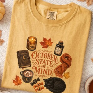 May include: A mustard yellow t-shirt featuring a graphic print with the text "OCTOBER STATE OF MIND". The design includes a lit candle, a spell book, a bottle labeled "witches brew", a mug of cocoa, a knit scarf, maple leaves, and a black cat.