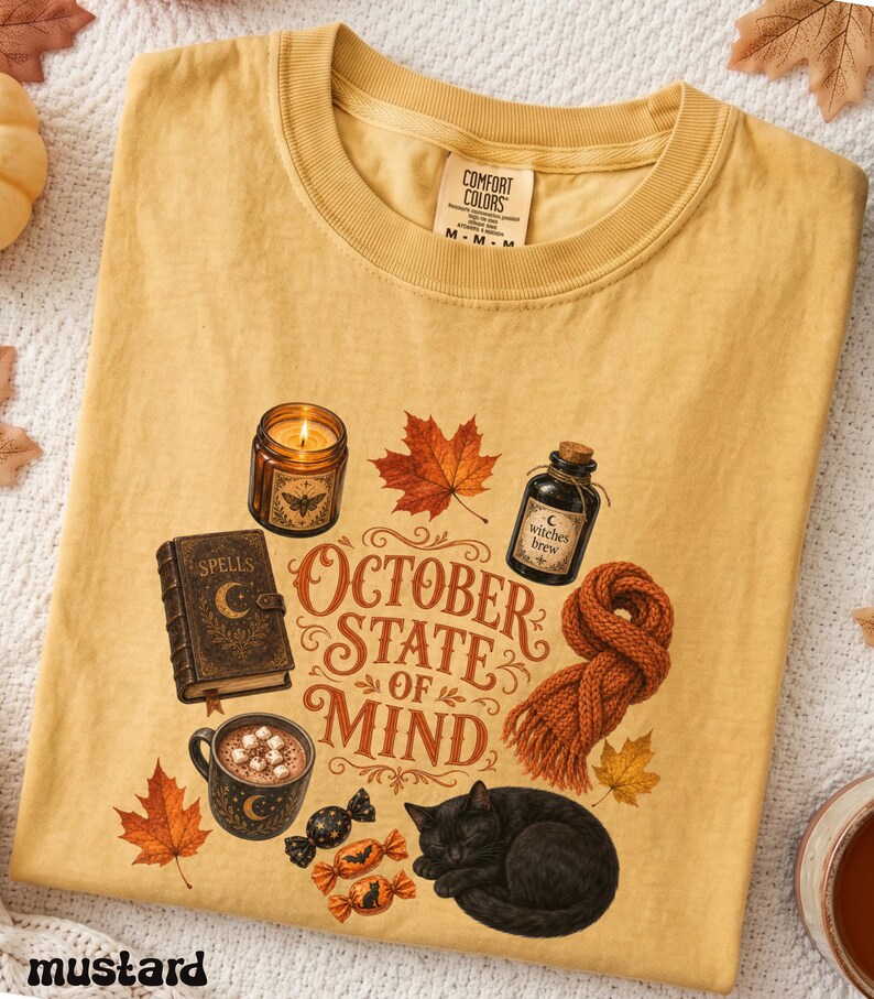 May include: A mustard yellow t-shirt featuring a graphic print with the text "OCTOBER STATE OF MIND". The design includes a lit candle, a spell book, a bottle labeled "witches brew", a mug of cocoa, a knit scarf, maple leaves, and a black cat.