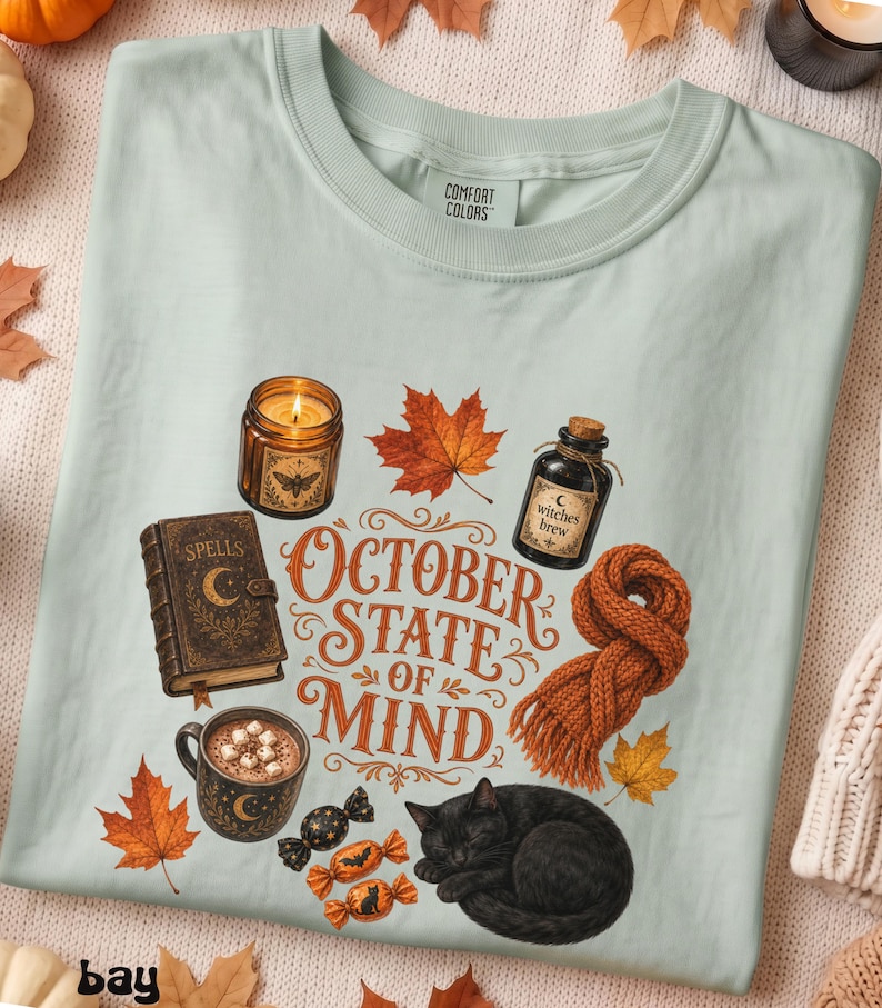 May include: A light sage green t-shirt featuring a graphic print with the text "OCTOBER STATE OF MIND". The design includes a lit candle, a spell book, a mug of cocoa, a black cat, a knitted orange scarf, autumn leaves, and wrapped candies.