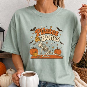 May include: A sage green t-shirt featuring a graphic of a skeleton performing a yoga pose on a mat. The text reads "Pilates & Bones" in orange and cream letters. The design includes orange pumpkins, black bats, and small stars surrounding the skeleton.