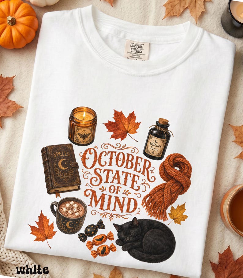 May include: A white t-shirt featuring a graphic print with the text "OCTOBER STATE OF MIND" surrounded by illustrations of a lit candle, a spell book, a bottle labeled "witches brew", a mug of cocoa, a knitted scarf, candies, and a black cat.