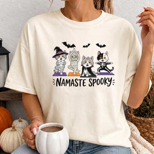 May include: An ivory t-shirt featuring a graphic of four cats performing yoga poses. The cats are dressed as a witch, a mummy, a vampire, and a skeleton. Above them are three black bats, and below the text reads NAMASTE SPOOKY in black letters.
