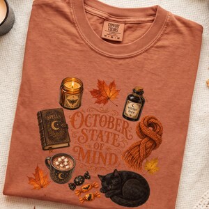 May include: A rust-colored t-shirt featuring a graphic print with the text "OCTOBER STATE OF MIND". The design includes a candle, a spell book, a bottle labeled "witches brew", a mug of cocoa, a knitted scarf, candies, and a black cat.