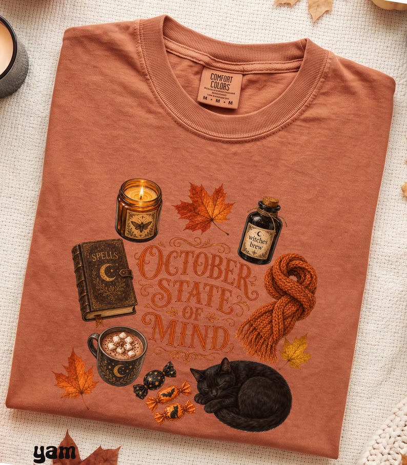 May include: A rust-colored t-shirt featuring a graphic print with the text "OCTOBER STATE OF MIND". The design includes a candle, a spell book, a bottle labeled "witches brew", a mug of cocoa, a knitted scarf, candies, and a black cat.