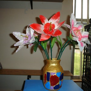 May include: A bouquet of artificial flowers in a gold vase with a mosaic design. The flowers are white, pink, and red lilies.