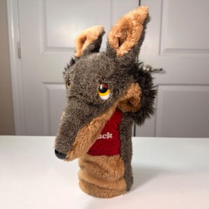May include: A plush hand puppet shaped like a wolf with dark gray and tan fur. The puppet features large pointed ears, yellow and black eyes, and a black nose. A red fabric panel on the chest displays the white text "ack" in a bold, sans-serif font.