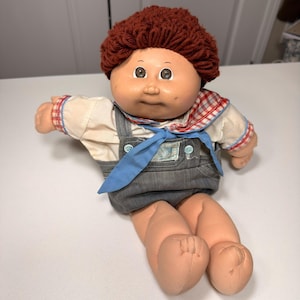 May include: A vintage Cabbage Patch Kid doll with a red yarn hairstyle. The doll wears a white shirt with red plaid trim, gray denim overalls, and a blue necktie. The doll has a peach-colored face and limbs.