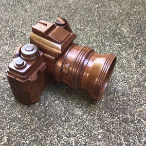 May include: A wooden camera model with a brown finish. The camera has a lens, a viewfinder, and buttons.
