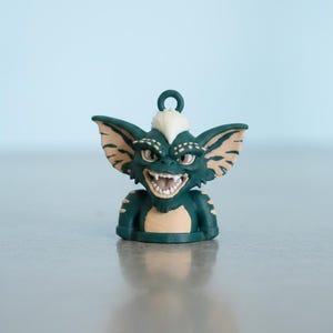 May include: A small, dark green creature figurine with large pointed ears, a white tuft of hair on its head, and a wide, toothy grin. The character has tan markings on its ears, chest, and shoulders, and features a small loop at the top for hanging.