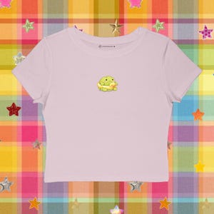 May include: A light pink crop top featuring a small graphic of a green frog hugging a yellow duckling. The shirt has a crew neck and short sleeves. The text "@cutetees.♥" is printed on the shirt.