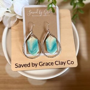 May include: Dangle earrings featuring a silver-toned hammered teardrop frame surrounding a hexagonal pendant with a marbled turquoise and white pattern. The earrings are displayed on a branded card labeled Saved by Grace Handmade Jewelry.