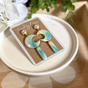 May include: A pair of dangle earrings featuring a white pearl stud attached to a crescent moon shape with a marbled turquoise and white pattern, finished with a matching rectangular drop. The earrings are displayed on a card labeled Saved by Grace.