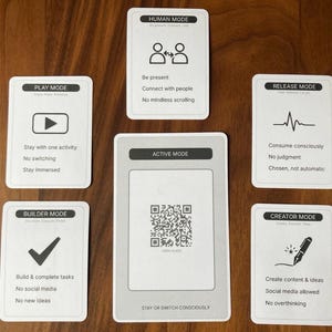 Può includere: Un insieme di carte bianche con testo e icone nere, disposte su una superficie di legno scuro. Le carte sono etichettate con termini come "PLAY MODE", "HUMAN MODE" e "CREATOR MODE". Ogni carta presenta una breve descrizione della sua modalità, incoraggiando la vita consapevole.