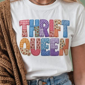 Peut inclure: T-shirt blanc avec l'inscription "THRIFT QUEEN" en lettres patchwork colorées. Les lettres sont composées de divers tissus à motifs, dont du vichy, des motifs floraux et des couleurs unies, cousus ensemble avec un fil contrastant.
