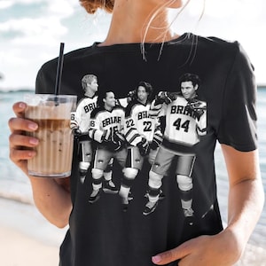 Op de afbeelding: Een zwart T-shirt met korte mouwen en een zwart-witte grafische print van vier hockeyspelers die truien dragen met de tekst BRIAR en verschillende nummers, waaronder 16, 22 en 44. De spelers zijn afgebeeld in volledige hockeyuitrusting.