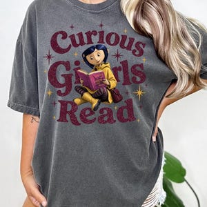 Pode incluir: Camiseta cinza escuro com o texto "Curious Girls Read" em bord&ocirc;. O gr&aacute;fico apresenta uma menina de desenho animado lendo um livro. A menina est&aacute; usando um casaco e botas amarelas. A camisa tem um visual vintage.