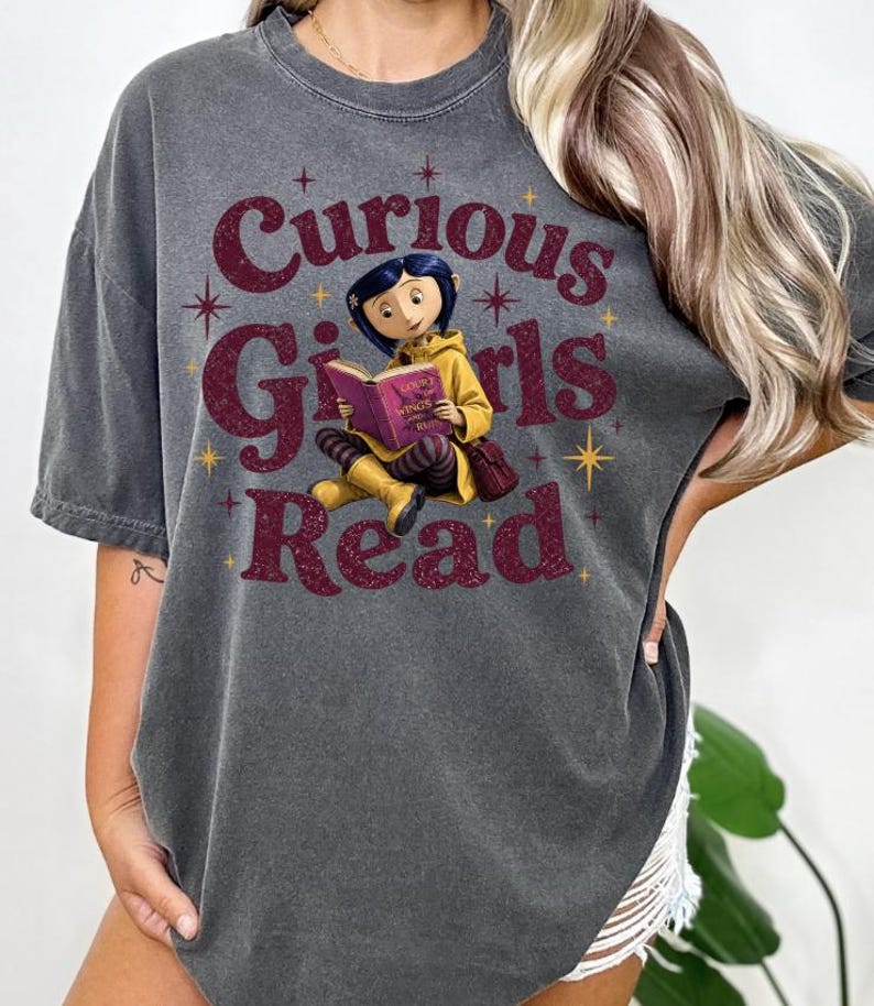 Pode incluir: Camiseta cinza escuro com o texto "Curious Girls Read" em bord&ocirc;. O gr&aacute;fico apresenta uma menina de desenho animado lendo um livro. A menina est&aacute; usando um casaco e botas amarelas. A camisa tem um visual vintage.