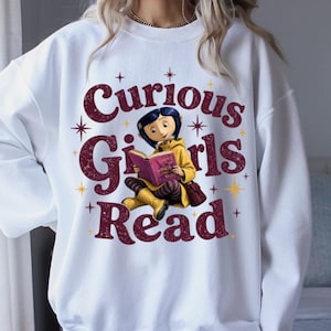 Pode incluir: Camisola branca com o texto "Curious Girls Read" em bord&ocirc;, apresentando uma menina de desenho animado lendo um livro. A menina est&aacute; usando um casaco e botas amarelas, com uma bolsa bord&ocirc;. O design inclui estrelas.