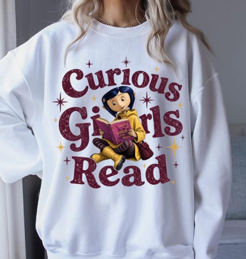 Pode incluir: Camisola branca com o texto "Curious Girls Read" em bord&ocirc;, apresentando uma menina de desenho animado lendo um livro. A menina est&aacute; usando um casaco e botas amarelas, com uma bolsa bord&ocirc;. O design inclui estrelas.