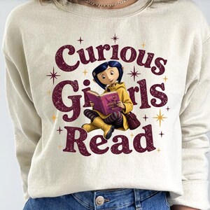 Pode incluir: Camisola creme com o texto "Curious Girls Read" em bord&ocirc;, apresentando um gr&aacute;fico de uma menina lendo um livro. A menina est&aacute; usando um casaco amarelo e sentada de pernas cruzadas. Explos&otilde;es de estrelas douradas acentuam o design.