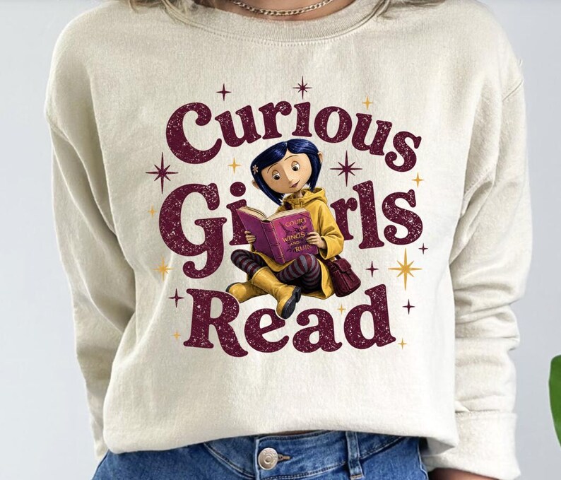 Pode incluir: Camisola creme com o texto "Curious Girls Read" em bord&ocirc;, apresentando um gr&aacute;fico de uma menina lendo um livro. A menina est&aacute; usando um casaco amarelo e sentada de pernas cruzadas. Explos&otilde;es de estrelas douradas acentuam o design.