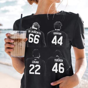Op de afbeelding: Een zwart t-shirt met korte mouwen en een witte grafische print van vier personen van achteren gezien, elk met een shirt met een naam en nummer: DI LAURENTIS 66, GRAHAM 44, LOGAN 22 en TUCKER 46, gerangschikt in een twee-bij-twee raster.