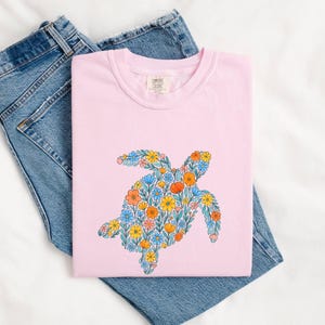 May include: A light pink short-sleeved t-shirt featuring a graphic of a sea turtle filled with a colourful pattern of orange, yellow, and blue flowers and green leaves. The shirt has a Comfort Colors brand label at the inner neckline.